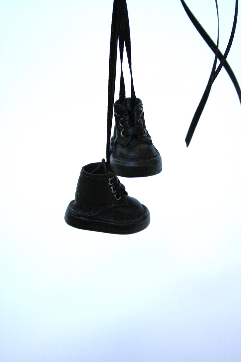 May include: Two miniature black leather boots hanging from black ribbons. The boots have laces and a classic design, with visible stitching details. The background is a plain white.