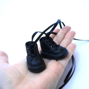 May include: A pair of miniature black leather boots with laces and long black ribbon ties. The small boots are held in a person's hand against a white background. The boots have a classic design.