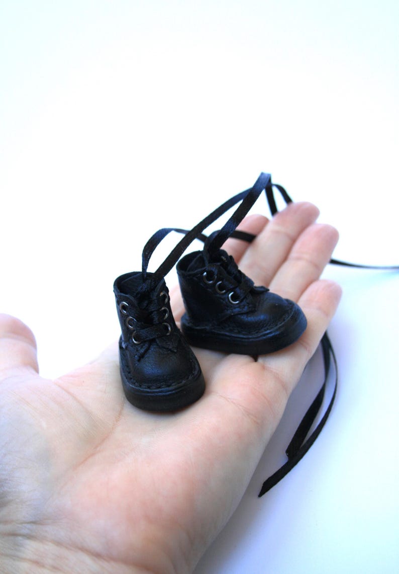 May include: A pair of miniature black leather boots with laces and long black ribbon ties. The small boots are held in a person's hand against a white background. The boots have a classic design.