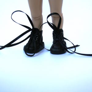 May include: A pair of small, black high-top sneakers with long, black laces. The shoes are laced up and tied, with the laces extending out to the sides. The sneakers are on a white surface.