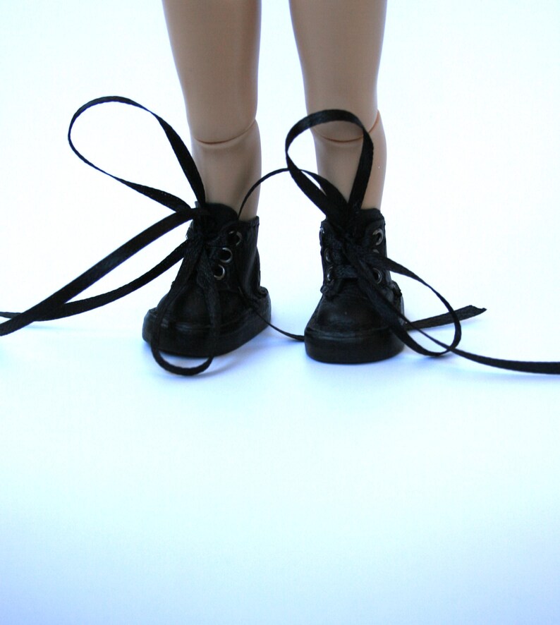 May include: A pair of small, black high-top sneakers with long, black laces. The shoes are laced up and tied, with the laces extending out to the sides. The sneakers are on a white surface.