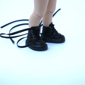 May include: A pair of black doll boots with laces. The boots are made of a smooth, dark material and have silver eyelets. The laces are long and extend beyond the boots. The boots are on a light background.