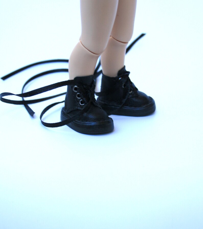 May include: A pair of black doll boots with laces. The boots are made of a smooth, dark material and have silver eyelets. The laces are long and extend beyond the boots. The boots are on a light background.
