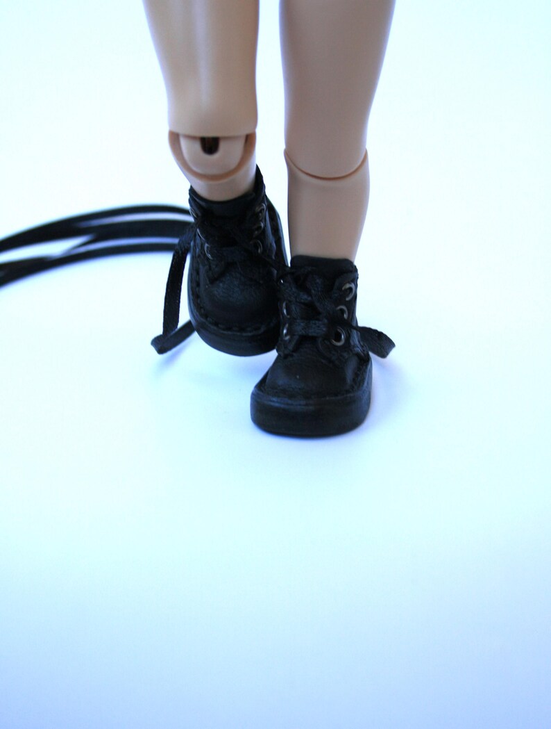 May include: Close-up of a pair of black, miniature lace-up boots. The boots are made of a textured material and have laces. The boots are on a light background. The boots are likely for a doll or other small figure.