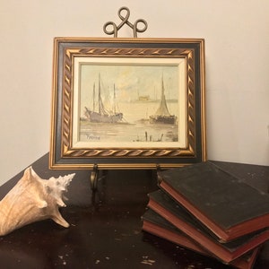Puede incluir: Una pintura enmarcada de un paisaje marítimo con dos veleros en el agua. La pintura está en un soporte dorado y hay una concha y tres libros sobre una superficie de madera oscura.