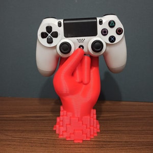 Può includere: Un controller bianco per PlayStation 4 è appoggiato su un supporto a forma di mano rosso brillante. Il supporto ha una base pixelata. Il controller presenta pulsanti neri e pad direzionali. Lo sfondo è blu-grigio tenue.
