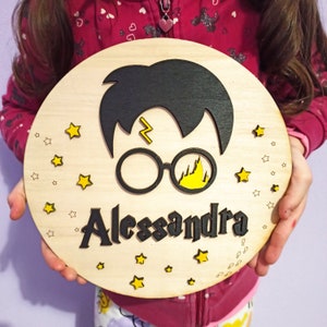 Può includere: Un cartello rotondo in legno con una silhouette nera di Harry Potter che indossa gli occhiali e una cicatrice a forma di fulmine. Il nome "Alessandra" è scritto in lettere nere sotto la silhouette. Il cartello è decorato con stelle gialle.
