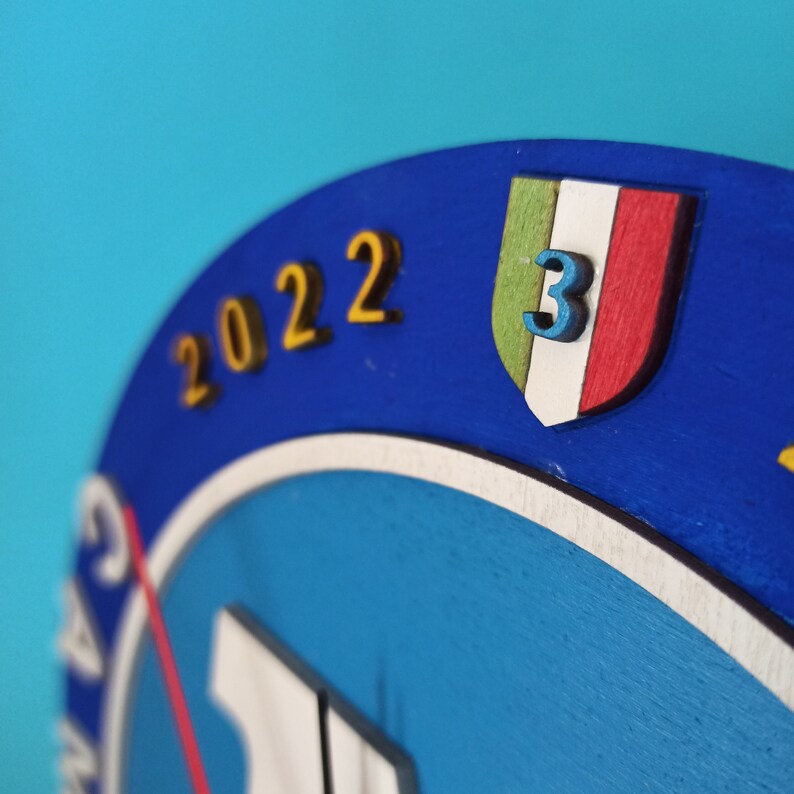 May include: A blue wooden clock face with the year "2022" in gold lettering. A shield with the Italian flag colors and the number "3" is featured. The clock has a white and red design.