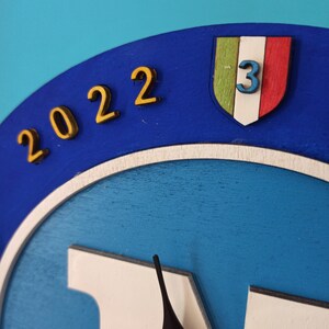 May include: A blue and white wooden clock with gold-colored numbers indicating the year 2022. The clock features a shield with the Italian flag colors and the number 3. The clock hands are black.