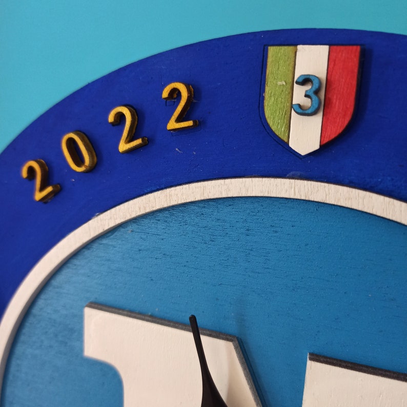 May include: A blue and white wooden clock with gold-colored numbers indicating the year 2022. The clock features a shield with the Italian flag colors and the number 3. The clock hands are black.