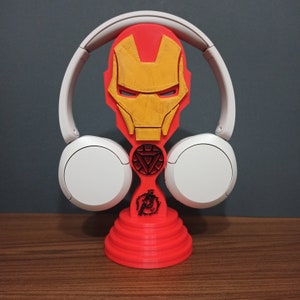 Può includere: Supporto per cuffie stampato in 3D rosso e giallo con una maschera di Iron Man stilizzata. Il supporto ha un simbolo di Iron Man nero sul davanti.
