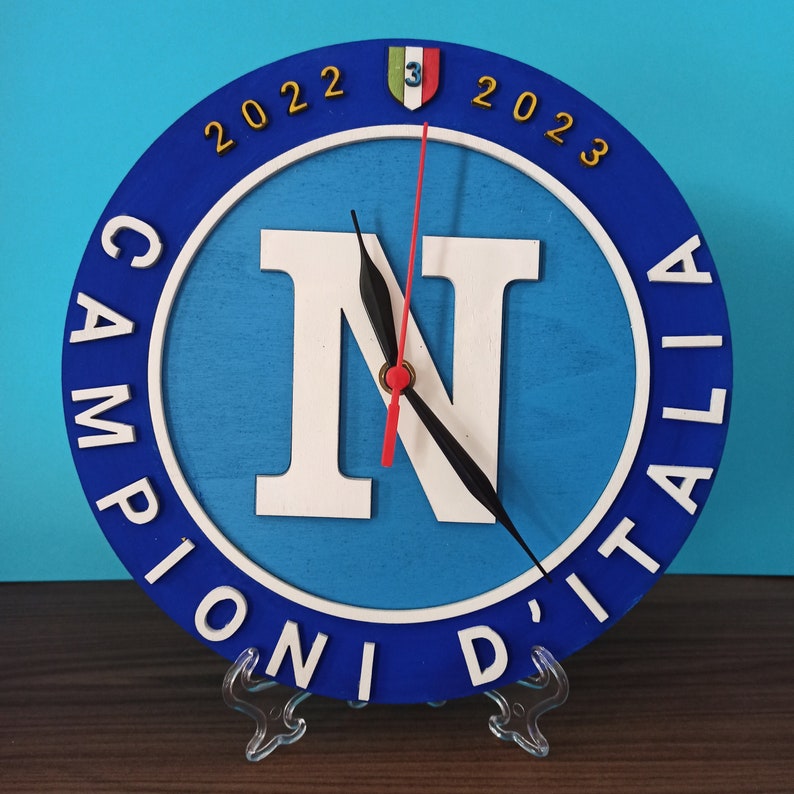 May include: A round, blue and white wooden clock with the text "CAMPIONI D'ITALIA" and the years "2022" and "2023". The clock face features a large white "N" and a red second hand.