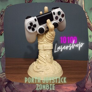 Op de afbeelding: Een witte videogamecontroller wordt vastgehouden door een zombiehand, die deel uitmaakt van een beige plastic standaard. De standaard heeft de tekst "PORTA JOYSTICK ZOMBIE" op de basis. De tekst "10100 Lasershop" is zichtbaar op de achtergrond.