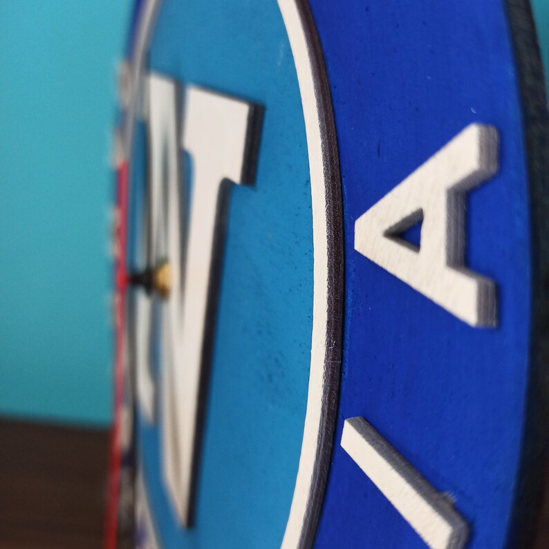 May include: A round wooden clock with a blue and white design. The clock face features large white letters against a blue background. The clock hands are visible, and the clock is mounted on a wall.