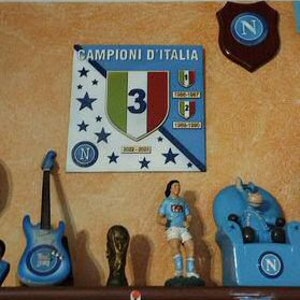 May include: A shelf displays various decorative items. A blue guitar, a trophy, a figurine in a soccer uniform, and a blue chair with the letter "N" are visible. A sign with the text "Campioni D'Italia" and a shield with the letter "N" are mounted on the wall.