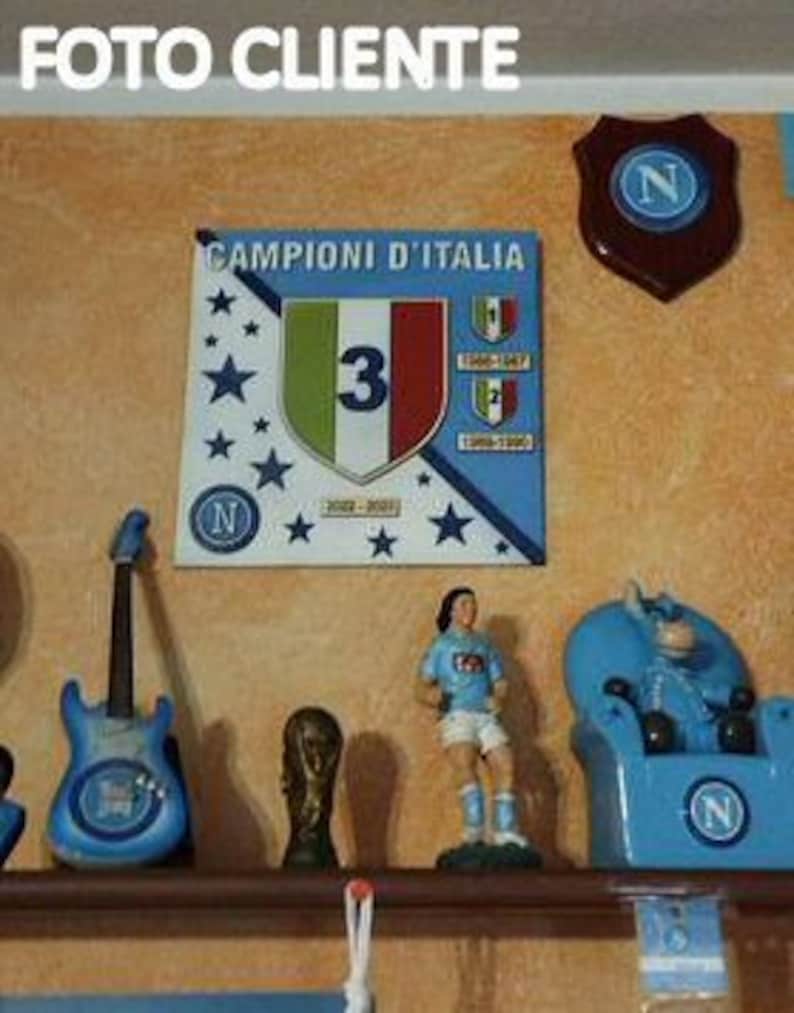 May include: A shelf displays various decorative items. A blue guitar, a trophy, a figurine in a soccer uniform, and a blue chair with the letter "N" are visible. A sign with the text "Campioni D'Italia" and a shield with the letter "N" are mounted on the wall.