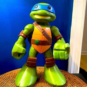 May include: A green Teenage Mutant Ninja Turtles action figure with a blue bandana and orange shell. The figure has a large head with a wide smile and is posed with its fists clenched.