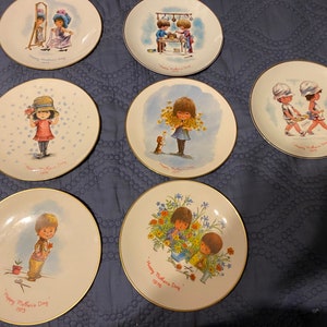 May include: Set of 7 white ceramic plates with gold trim. Each plate features a different illustration of children celebrating Mother's Day. The plates are decorated with flowers, hearts, and other festive designs. The text "Happy Mother's Day" is printed on each plate.