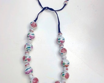 May include: A white beaded necklace with a dark blue cord. The beads are round and decorated with red Chinese characters, pink flowers, and green leaves. The necklace is on a white surface.