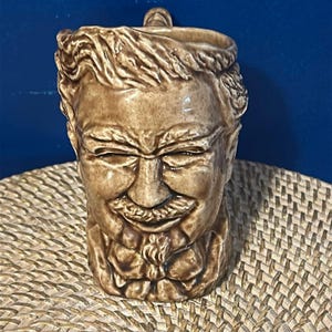 May include: A brown ceramic mug shaped like a man's head with a mustache and a goatee. The mug has a handle on the side and is decorated with details like hair and a bow tie.