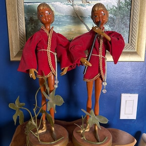 May include: Two tall, slender figurines with brown skin and blonde hair, each wearing a red velvet tunic with gold trim and a white cord belt. One figurine holds a black flute to its lips. Both stand on round wooden bases with green plastic vines.