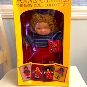 May include: A yellow box with a red and blue baby doll inside. The doll has blonde curly hair and is wearing a blue and green striped sweater and a red skirt. The box says "Anne Geddes The Baby Doll Collection" and "For ages over 18 months".