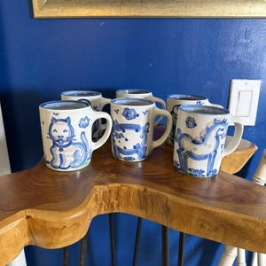 Könnte beinhalten: Ein Set aus fünf weißen Keramikbechern mit handgemalten blauen Illustrationen verschiedener Tiere, darunter eine Katze, ein Pferd und abstrakte Formen. Jeder Becher verfügt über einen geschwungenen Henkel und eine glatte Oberfläche.