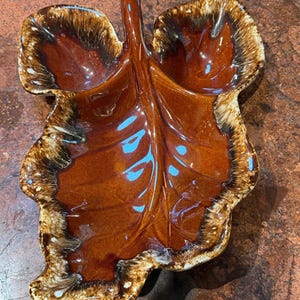 May include: A brown ceramic leaf-shaped dish with a glossy finish. The dish has a raised handle and a detailed leaf vein design. The edges are a darker brown with white and tan accents, creating a unique, decorative piece.