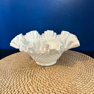 May include: A white milk glass bowl with a ruffled edge and a textured, dimpled surface. The bowl is sitting on a woven mat.