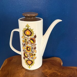 May include: A vintage white ceramic coffee pot with a brown lid and handle. The pot is decorated with a floral pattern in shades of yellow, orange, and brown. The pot is on a wooden surface.