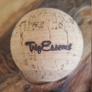 May include: A round cork bottle stopper with the word "Trig Essence" in black script. The stopper is made of light brown cork with a textured surface. The product is designed for sealing bottles.
