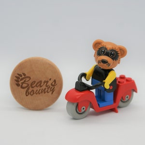 May include: A brown bear wearing a black shirt, yellow vest, and blue jeans rides a red toy motorcycle with black handlebars and gray wheels. The motorcycle has a red seat and a black exhaust pipe. A wooden round disc with the text "Bear's bounty" is in the background.