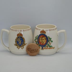 Może przedstawiać: Dwa białe ceramiczne kubki z motywem królewskim. Jeden kubek przedstawia herb królewski z literami "G.R." i tekstem "2nd MAY 1957 - CORONATION." Drugi kubek przedstawia portret królowej Elżbiety II i księcia Filipa z tekstem "QUEEN ELIZABETH II" i "DIEU ET MON DROIT".