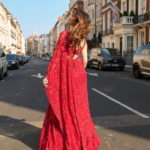 May include: A vibrant red sari with intricate embroidery and embellishments. The traditional Indian garment features a flowing skirt and a draped shawl, creating a graceful silhouette. The sari is worn outdoors on a city street.