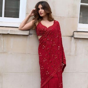 May include: A vibrant red saree with intricate embroidery and mirror work. The saree features a detailed bodice and a flowing drape, showcasing a rich texture and design. The garment is a traditional Indian outfit, perfect for special occasions.