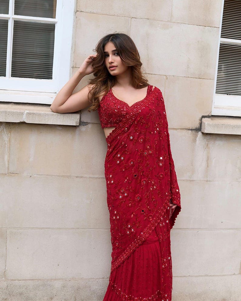 May include: A vibrant red saree with intricate embroidery and mirror work. The saree features a detailed bodice and a flowing drape, showcasing a rich texture and design. The garment is a traditional Indian outfit, perfect for special occasions.