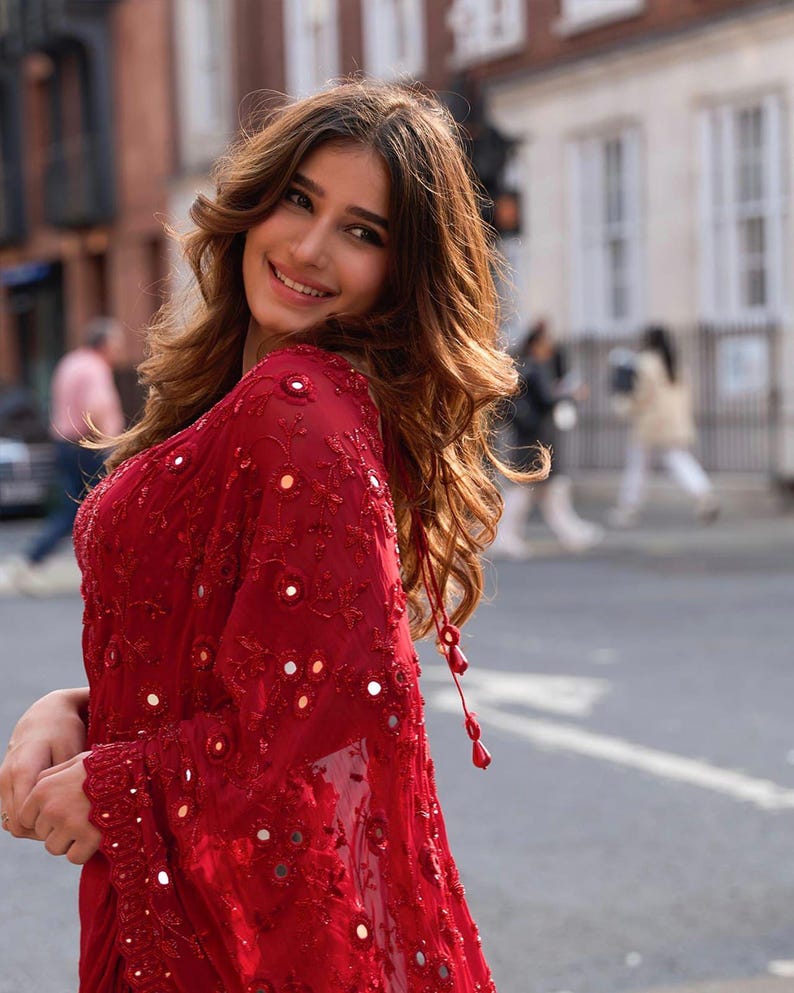 May include: A red sari with intricate embroidery and mirror embellishments. The sheer fabric drapes elegantly, with long sleeves and a decorative tassel. The sari is a vibrant shade of red, perfect for special occasions.