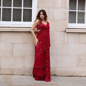 May include: A red formal dress with a V-neckline and intricate embellishments. The dress features a layered skirt and a draped design, creating an elegant silhouette. The fabric appears to have a subtle sheen.