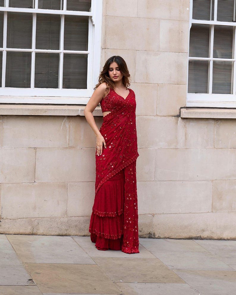 May include: A red formal dress with a V-neckline and intricate embellishments. The dress features a layered skirt and a draped design, creating an elegant silhouette. The fabric appears to have a subtle sheen.