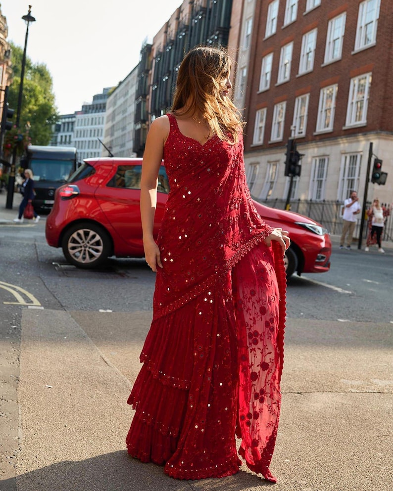 May include: A vibrant red saree with intricate sequin and bead embellishments. The saree features a layered skirt and a sheer, floral-patterned drape. The sleeveless top has a sweetheart neckline. The image is taken outdoors, with a red car and buildings in the background.