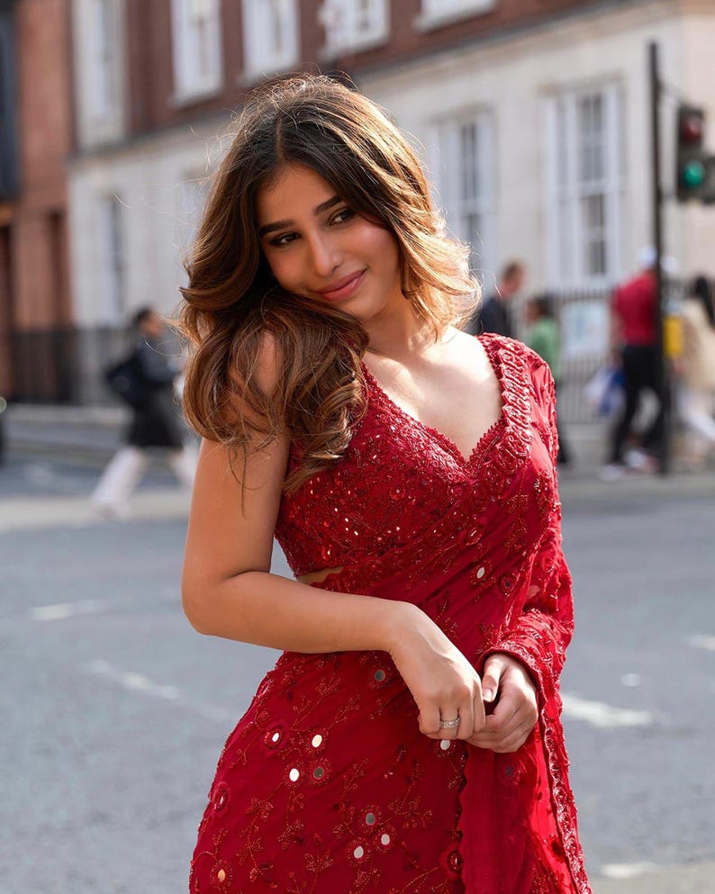 May include: A red sari with intricate embroidery and mirror embellishments. The sari features a V-neckline and long sleeves. The image shows a woman wearing the sari outdoors.