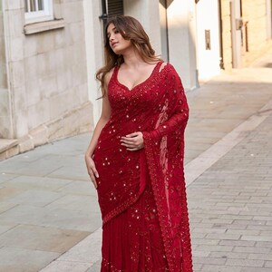 May include: A vibrant red saree with intricate sequin and bead embellishments. The saree features a plunging neckline and delicate shoulder straps. The fabric drapes elegantly, showcasing a flowing design, perfect for special occasions.