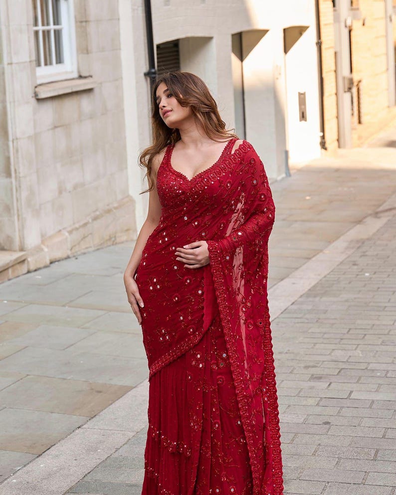 May include: A vibrant red saree with intricate sequin and bead embellishments. The saree features a plunging neckline and delicate shoulder straps. The fabric drapes elegantly, showcasing a flowing design, perfect for special occasions.