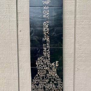 May include: Black wooden sign with gold lettering. The sign has a poem written on it that reads: "Smell like teen spirit on a plain come as you are you know you're right been a son paper cuts: lithium something in the way about a girl all apologies in bloom drain you lounge act breed the man who sold the world where did you sleep last night"