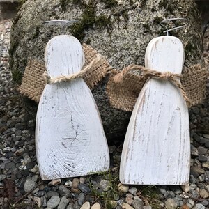 May include: Two white wooden angels with burlap wings and wire halos. The angels are standing on a bed of small gray rocks.