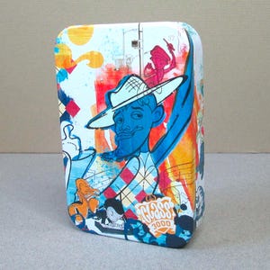 May include: A rectangular tin with a colorful, cartoonish design. The artwork features a blue-skinned man in a white hat, a blue arm, and a diamond-patterned shirt. The tin has a white background with orange, red, and blue accents. The text "GLASS 3000" is visible.