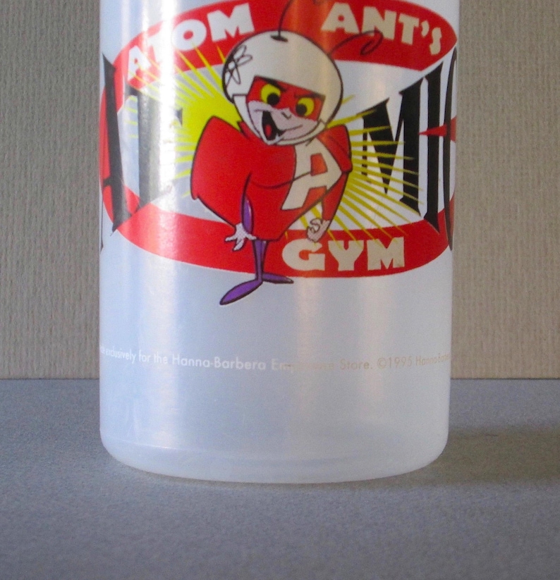 Puede incluir: Una botella de agua de pl&aacute;stico transparente con un logotipo de Atom Ant's Gym rojo, amarillo y negro. El logotipo presenta una hormiga de dibujos animados que lleva un casco y un traje rojo con una gran letra "A" en el pecho.