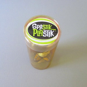 K&ouml;nnte beinhalten: Ein durchsichtiger Plastikbeh&auml;lter mit einem gelben und gr&uuml;nen Deckel. Der Deckel tr&auml;gt den Text "SPASTIK PLASTIK" in schwarzen Buchstaben auf wei&szlig;em Hintergrund.