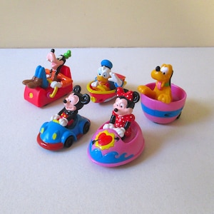 Op de afbeelding: Een set van vijf kleurrijke plastic Disney-speelgoedfiguren. Mickey Mouse, Minnie Mouse, Donald Duck, Goofy en Pluto rijden elk in een ander voertuig. Het speelgoed is in uitstekende staat.