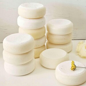 May include: Several stacks of small, round, off-white objects, possibly soap or bath bombs, are arranged on a white surface. Some stacks are taller than others. A small yellow flower rests on top of one of the objects.